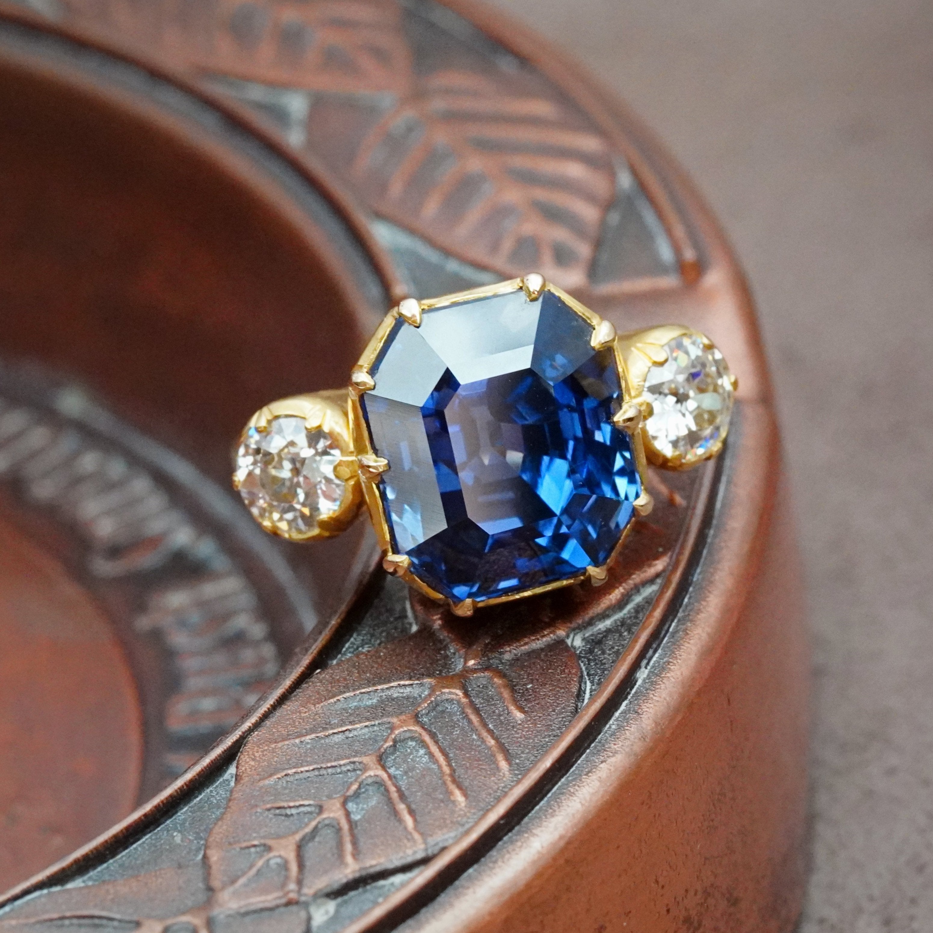 An exquisite 12.81 ct step-cut Madagascar sapphire, no heat, radiating vivid blue brilliance. Set in a 22K yellow gold prong setting with diamond accents, available at Jogani.